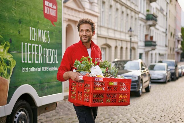 A REWE delivery service employee carrying a red crate with groceries. In the background is his delivery van. | © REWE