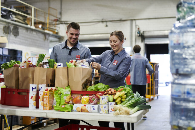 Two REWE delivery service employees packing fresh groceries into bags in the warehouse. | © REWE