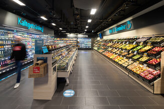 Innenansicht eines modernen REWE Markts mit Abteilungen für Kaltgetränke und Obst & Gemüse, ein Kunde geht durch den Gang | © REWE
