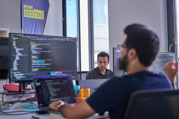 Zwei Männer arbeiten konzentriert an Computern, im Hintergrund ist ein REWE digital Banner zu sehen