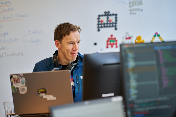 Ein Mitarbeiter von REWE digital arbeitet an seinem Laptop, mit Kopfhörern um den Hals und einem Lächeln auf dem Gesicht | © REWE digital, Sebastian Knoth