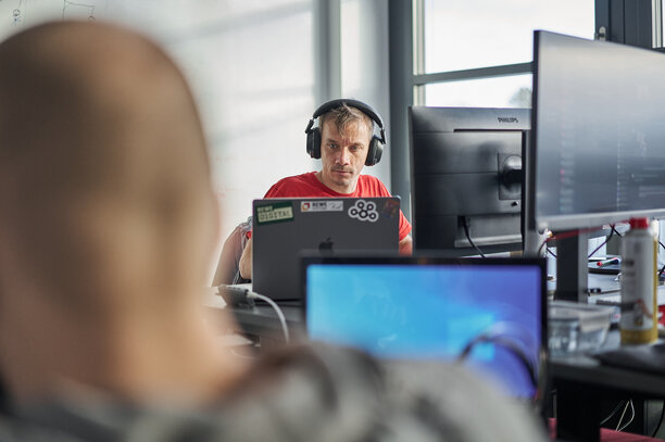 Ein Mann mit Kopfhörern arbeitet konzentriert an einem Schreibtisch mit mehreren Monitoren in einem modernen Büro | © REWE digital, Sebastian Knoth