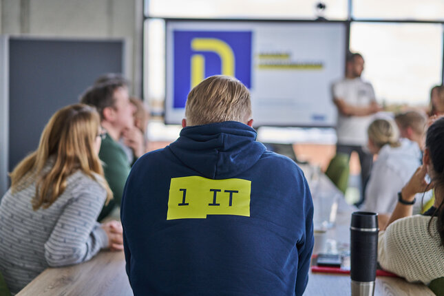 Eine Gruppe von Mitarbeitenden sitzt bei einer Präsentation im Besprechungsraum, im Vordergrund eine Person mit “1 IT”-Hoodie | © REWE digital, Sebastian Knoth