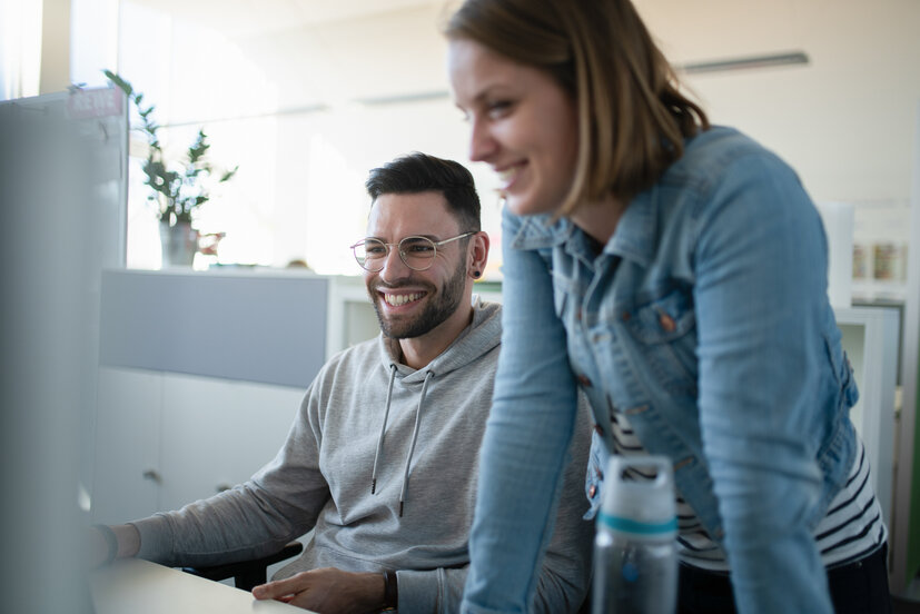 Zwei IT-Expert:innen von REWE digital arbeiten zusammen an einem Computerbildschirm in einem Büro.