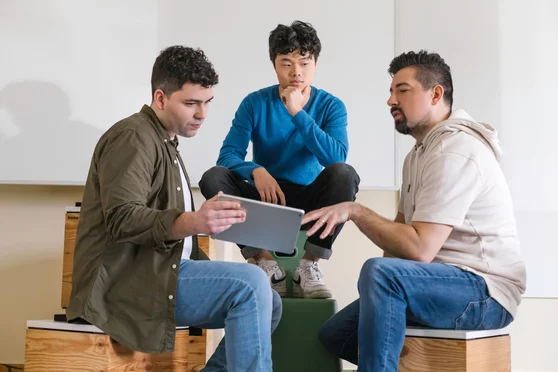 Drei Personen sitzen auf Hockern in einem Büro und schauen gemeinsam auf ein Tablet, während sie miteinander arbeiten und sich austauschen. | © REWE digital, Paula Winkler und Alexander Gehring