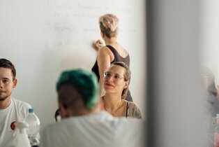 Eine Gruppe diverser Mitarbeiter:innen arbeitet an einem Tisch zusammen, eine Frau schreibt im Hintergrund etwas ans Whiteboard | © REWE digital