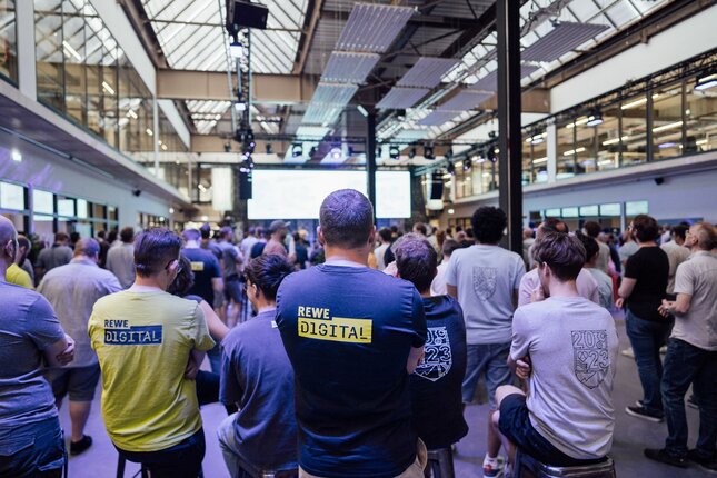 Mitarbeitende sind von hinten zu sehen, wie sie bei einem Firmenevent auf eine Leinwand schauen | © REWE digital, hunderteins GmbH