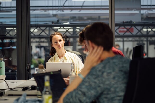 Eine IT-Expertin sitzt am Schreibtisch und schaut zu zwei Kollegen herüber | © REWE digital