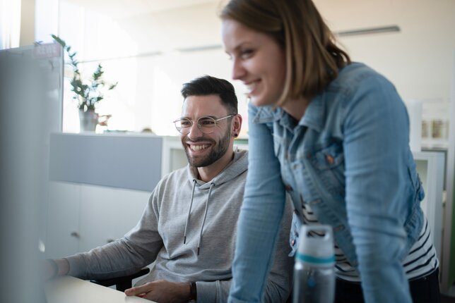 Two employees smiling and working together on a computer in the office | © REWE digital, hunderteins GmbH