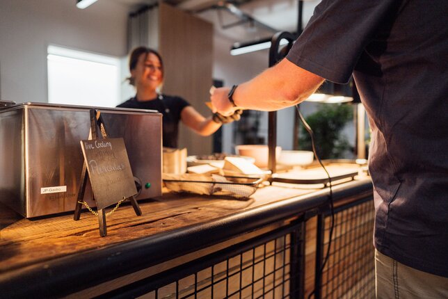 Symbolic image for permanent discounts: an office kitchen employee handing a cup of coffee to a colleague. | © REWE digital, hunderteins GmbH