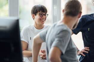 Eine IT-Expertin lächelt beim Gespräch mit Kolleg:innen im Büro | © REWE digital