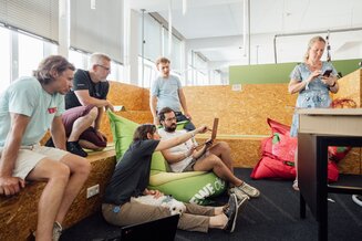Mehrere IT-Expert:innen sitzen in einem modernen Büro auf Sitzsäcken und Holzbänken und arbeiten gemeinsam an einem Laptop | © REWE digital