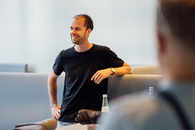 Ein stehender Mann in einem schwarzen T-Shirt stützt einen Arm auf einen Computermonitor und lächelt.  | © REWE digital