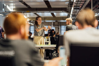 Person präsentiert Ideen vor einer Gruppe in einem modernen Büro. Im Vordergrund sind unscharf weitere Teilnehmende zu sehen. Das Bild zeigt einen Workshop, Austausch und kollaboratives Arbeiten während eines REWE digital Hack-Events. | © REWE digital