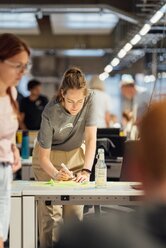 Teilnehmende arbeiten in einem modernen Workshop-Bereich zusammen. Im Mittelpunkt notiert eine Person Ideen auf Haftnotizen an einem Tisch, während weitere Teilnehmende im Raum an ihren Projekten arbeiten. Das Bild zeigt eine kollaborative Arbeitsphase während eines REWE digital Hack-Events. | © REWE digital, hunderteins 