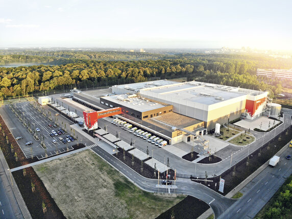 Luftaufnahme eines modernen Logistikzentrums der REWE Group. Das weitläufige Gelände umfasst Lagerhallen, Lieferfahrzeuge, Parkflächen und Zufahrtsstraßen. Das Bild zeigt die logistische Infrastruktur für die Versorgung von Märkten und Kund:innen. | © REWE Group