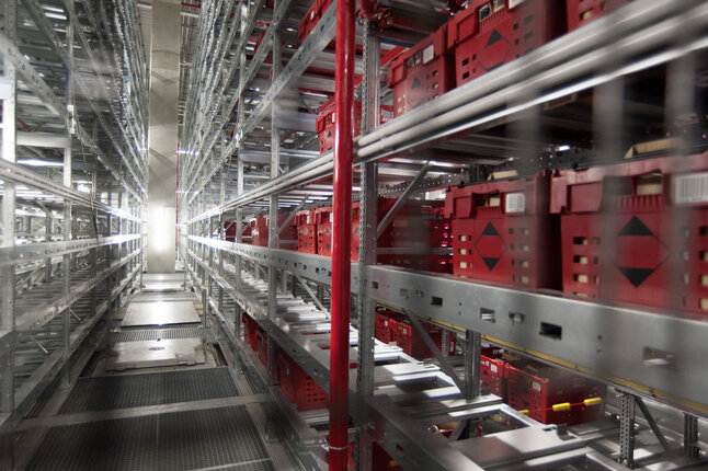 Regalanlage eines automatisierten Logistiksystems mit zahlreichen roten Transportbehältern. Die Behälter bewegen sich durch ein mehrstöckiges Lager aus Metallregalen und Förderelementen. Das Bild zeigt die technologische Infrastruktur für effiziente Warenlagerung und Kommissionierung bei REWE. | © REWE Group