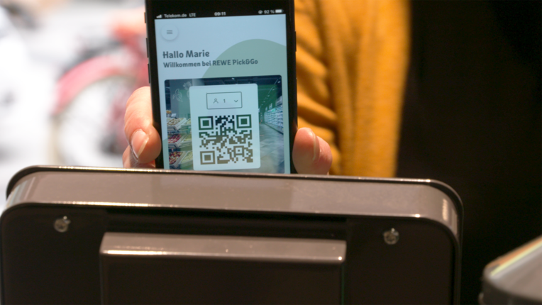 Smartphone mit geöffneter REWE Pick&Go App und QR-Code vor einem Scanner im Markt. Das Bild zeigt den digitalen Zugang zum kassenlosen Einkauf über die App. | © REWE