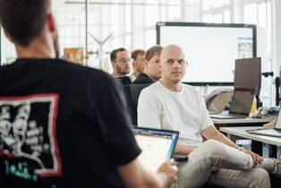 Mitarbeiter:innen sitzen mit Laptops in einem hellen, modernen Büroraum und hören aufmerksam einem Kollegen zu